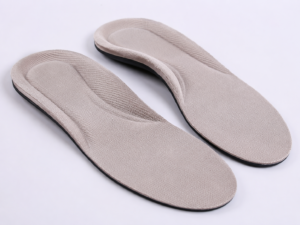 Realistic close-up of a pair of soft cushioned shoe insoles with textured beige fabric, contoured heel padding, and black edge trim on a clean white background.