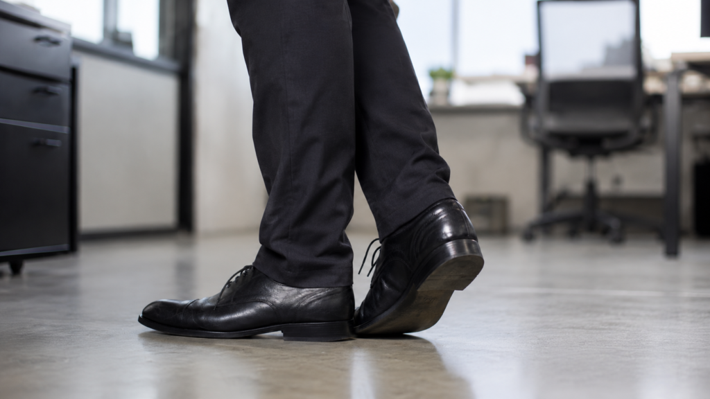 Close-up of a person in formal black shoes standing on a hard office floor, shifting weight from one foot to the other, showing discomfort from standing for long hours.